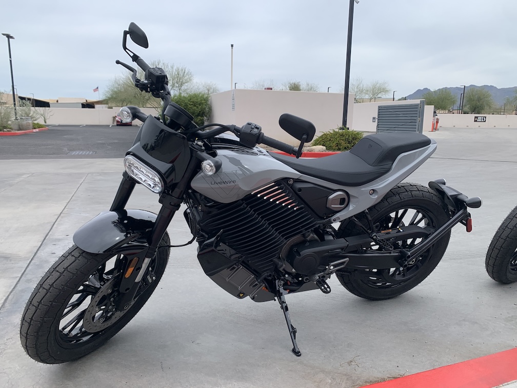 A gray and black motorcycle resting on its kickstand next to a red curb in a parking lot, on a cloudy day