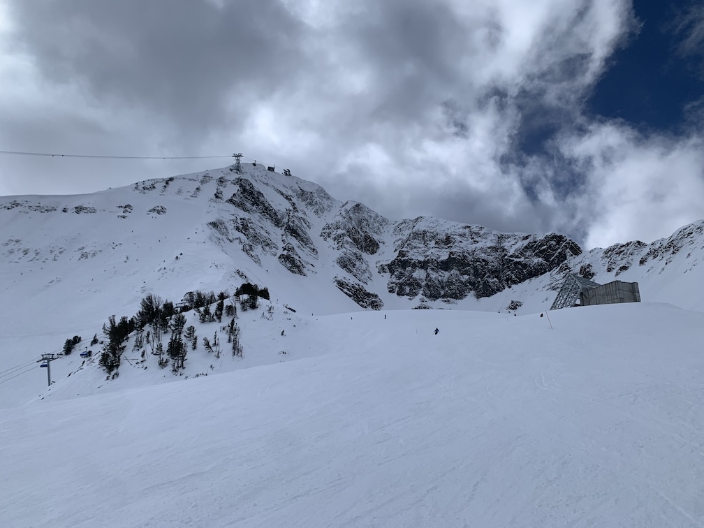 A snowy, rocky, steep mountain top on an overcast day, with a tram tower and structure at the top, and a few skiers visible visible in the midground