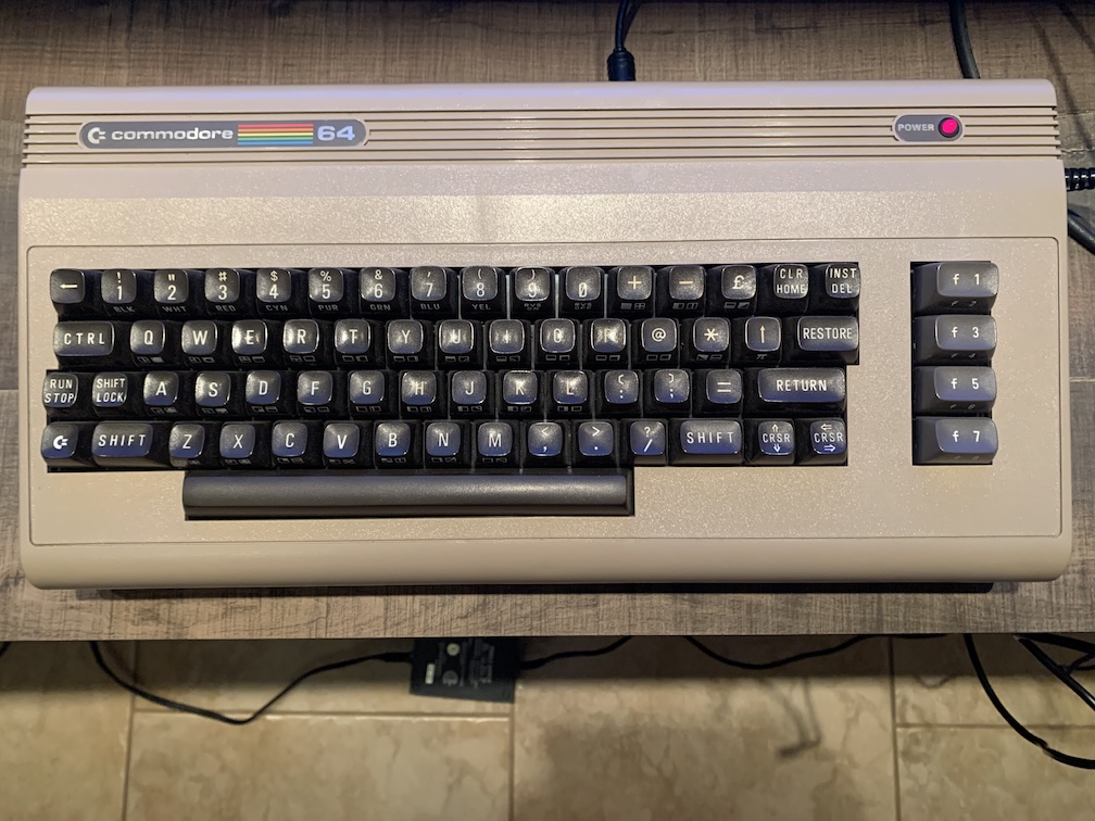 A computer sitting on a wooden surface above the floor, consisting of a dark beige rectangular case about twice the size of the type-writer style keyboard, with four large keys to the right of the keyboard and a red LED in the upper right-hand corner and the computer's model name in the upper left-hand corner