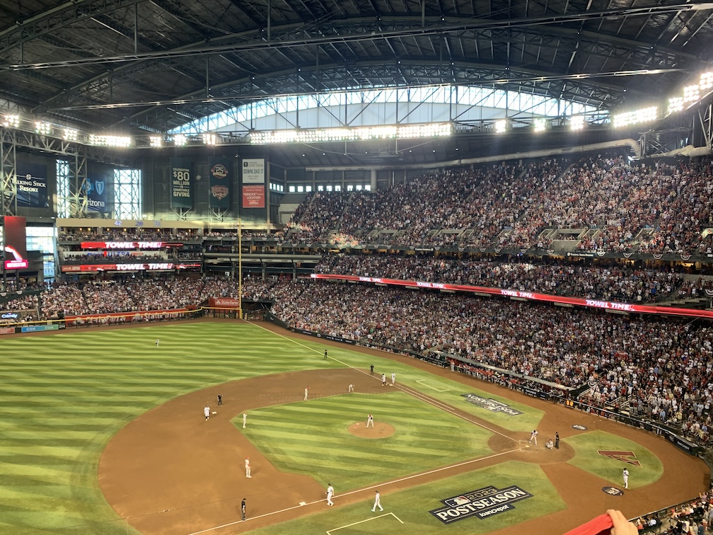 A view of a baseball diamond and the right field portion of the outfield, taken from the upper deck of the third base stands, with defenders in grey uniforms on the dirt of the infield and runners in white uniforms on all three bases and a leftie batter at the plate, beyond which are the stands of the stadium which are packed with fans, and above which is a large arched bank of windows and a metal roof