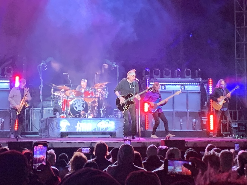 Five musicians on a stage with a drumset and speakers in the background and people's heads below in the foreground: a saxophone player, a drummer, a guitarist dressed in black, a bass player, and another guitarist