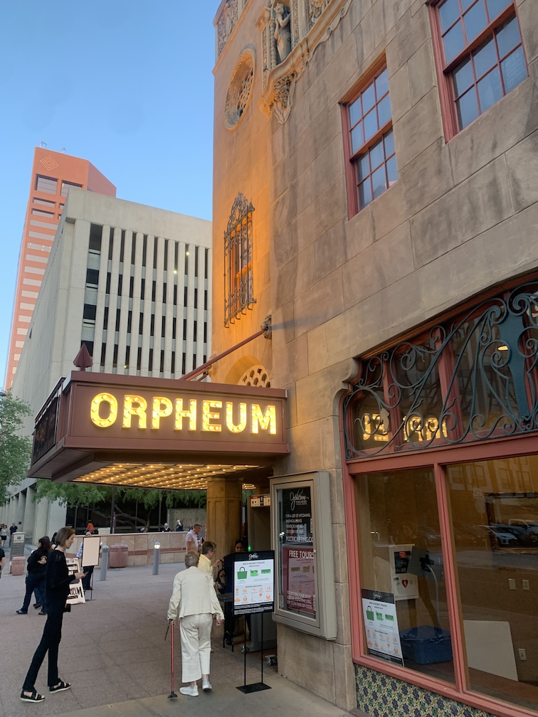 The exterior of one side of a theater, with a lighted marquee featuring the theater's name hanging on the side of a stone building with some windows and intricate carvings, with a few people in front about to enter the theater and some modern office buildings in the background