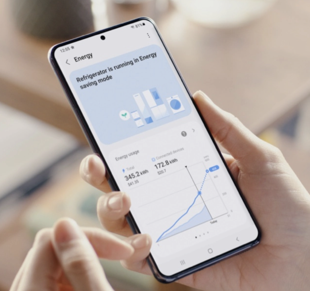 A smartphone held in a hand, the screen of which shows that the refrigerator is running in 'Energy saving mode' above a graph of the energy usage in units of kilowatt-hours